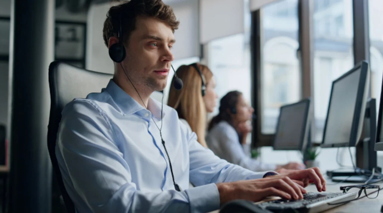 A man seen from the side, sitting at a desk with a headset on, typing on his PC.