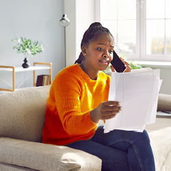 Eine Frau mit ein paar Seiten Papier in der Hand, die auf einem Sofa sitzt und mit besorgtem Gesicht telefoniert