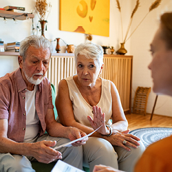Zwei &auml;ltere Personen, die einer Frau, die nur halb im Bild ist, gegen&uuml;bersitzen und etwas erkl&auml;ren.