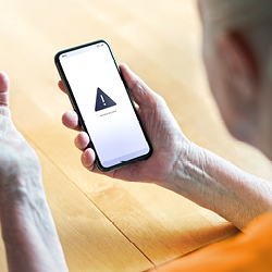 Eine Person hält ein Smartphone in der Hand, auf dessen Bildschirm ein Warnsymbol zu sehen ist