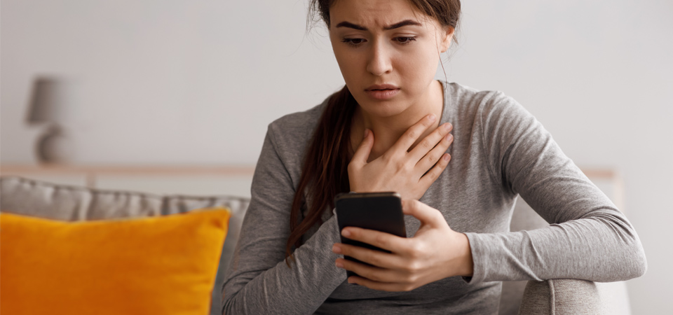 A woman sitting on a sofa, clutching her chest with a distressed expression while looking at her smartphone