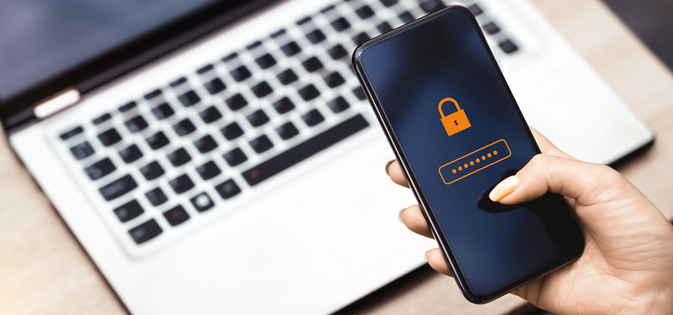 A hand holding a smartphone displaying a lock symbol and an anonymized PIN, with an open laptop in the background